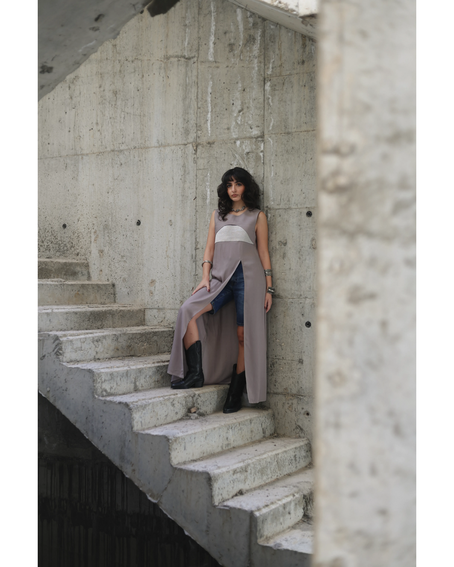 Farah Daylac warm grey sleeveless maxi dress with embroidered chest band and deep front slit, worn over jeans.